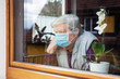 © M.Dörr & M.Frommherz - Senior woman with surgical mask sitting on a window at home, coronavirus and covid-19 provisions