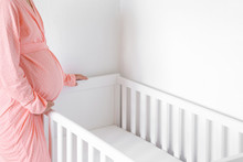 One Young Pregnant Woman In Pink Robe Standing Beside White Crib For Future Baby. Waiting Concept. Closeup. Side View.