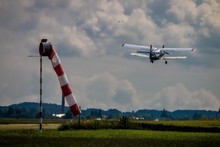 Photo Of A Small Field Airport With Red And White Windsock And A Biplane Taking Off. Green Field And Summer Clouds In The Background.