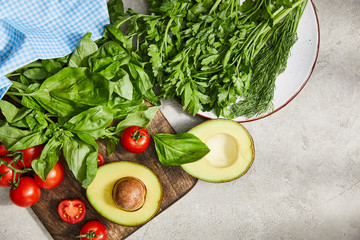 Canvas Print - Top view of fabric near plate with greenery and cutting board with cherry tomatoes, basil leaves and avocado halves on grey