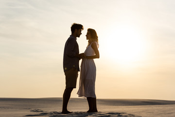 Wall Mural - side view of young couple holding hands on sandy beach at sunset