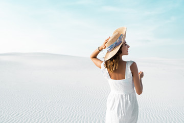 back view of beautiful girl in white dress and straw hat on sandy beach with blue sky
