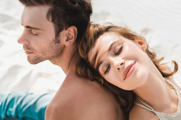 Canvas Print - smiling young couple sitting back to back on beach