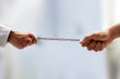 © mirzamlk - Shot of two hands on the end of a rusted metal chain pulling from both sides with blurred background and selective focus. Male hand playing tug of the war with a metal chain.