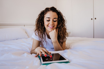 .Young caucasian woman lying on the bed in her room using her tablet to communicate with her family during the quarantine caused by covid19. Lifestyle