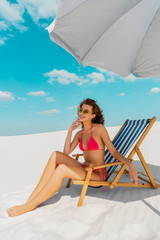 Wall Mural - smiling beautiful sexy girl in swimsuit and sunglasses sitting in deck chair under umbrella on sandy beach