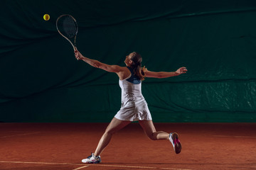 Wall Mural - Young caucasian professional sportswoman playing tennis on sport court background. Training, practicing in motion, action. Power and energy. Movement, ad, sport, healthy lifestyle concept. Front view.