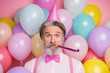 Close-up portrait of his he nice attractive cheerful funky grey-haired mature man having fun blowing festal whistle among colorful air balls isolated over pink pastel color background