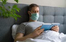 Sick Man With Medical Mask Reading Book In Bed. Home Quarantine Concept.