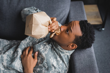 Wall Mural - stressed african american soldier in military uniform breathing with paper bag while having panic attack and suffering from PTSD at home