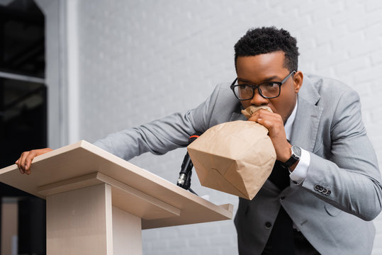 stressed african american speaker breathing with paper bag and having panic attack on business confe