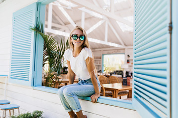 Interested woman in stylish sparkle sunglasses sitting on window sill. Photo of gorgeous blonde female model with suntan posing with sincere smile.