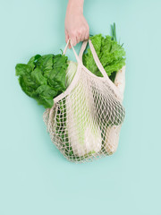 The hand holds an eco bag with healthy vegetables. Cabbage, spinach leaves, lettuce, onion on a mint background. Eco Shopping.