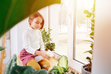 Wall Mural - Image of a little girl shot through leaves sitting on a sill, spraying seedlings