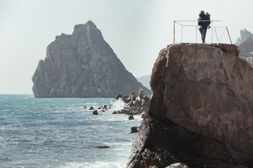 A woman with a small child in her arms stands on the edge of a large stone and looks at the stormy sea.