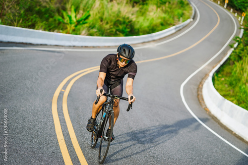 asian male riding on a black bicycle up and down the hill on the road, smiling and wearing bicycle crash helmet and goggles, on a long winding road with barriers and forest trees at the background.