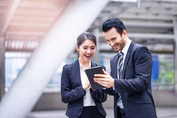 caucasian businessman holding a smart tablet device touching on the screen, showing information to asian businesswoman, outdoor in the city district with urban architectural structure in background