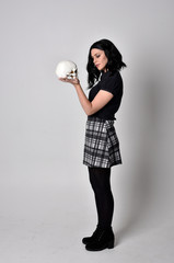  Portrait of a goth girl with dark hair wearing blue and plaid skirt with boots. Full length standing pose, holding a human skull, on a studio background.