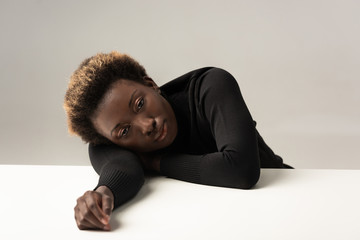 Wall Mural - tired african american woman in black turtleneck sitting lying on table isolated on grey