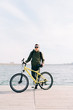 © Дарья Фомина - a young man in khaki sportswear and glasses stands on a pontoon near the lake and holds a yellow Bicycle near him on a Sunny spring day