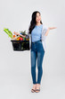 © Atstock Productions - Beautiful Asian woman with open hand holding supermarket shopping basket full of vegetables and groceries isolated on studio light gray background
