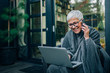 © bnenin - Smiling elder woman using laptop and talking on mobile phone, portrait.