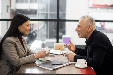 Wall Mural - side view of businessman and smiling asian businesswoman talking in cafe