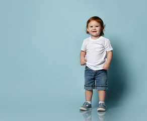 Little ginger toddler male or female in white t-shirt, socks and shoes, denim shorts. Child is smiling, posing on blue background