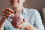 Fototapeta  - female hands hold a pink piggy bank and puts a coin there. The concept of saving money or savings, investment