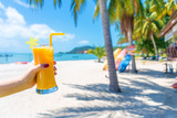 Fototapeta Panele - First-person view. Girl holds a glass cup of cold mango fresh on the background of a sandy tropical beach. White sand and palm trees. Fairytale vacation