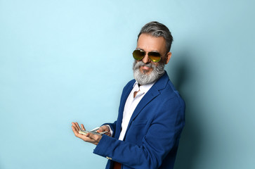 Wall Mural - Elderly man in white shirt, jacket, brown pants and sunglasses. He showing fan of hundred dollar bills, posing on blue background