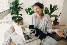 Cute Cat Helping Owner During Quarantine, Loyal Companion. Casual Girl Working On Laptop With Her Cat, Sitting Together In Modern Room With Pillows And Plants. Home Office.  Stay Home Stay Safe