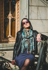 Wall Mural - Fashion, brunette woman in a dress and leather black jacket sitting on old iron chair outdoors. Girl model posingl in Batumi, Georgia. Old city at background.