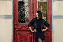 Brunette Woman Wearing In Black Leather Jacket Standing Near Old Red Door. Rock Concept. Girl Model In The Batumi, Georgia.