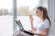 © blinova - Home quarantine.Protection from coronavirus. Caucasian woman at the window with a laptop. The girl waves her hand in greeting and smiles happily. Communicates online with friends and relatives.