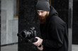 © Volodymyr - Bearded Professional videographer in black hoodie holding professional camera on 3-axis gimbal stabilizer. Filmmaker making a great video with a professional cinema camera. Cinematographer.