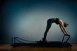 © Georgii - Young girl doing pilates exercises with a reformer bed. Beautiful slim fitness trainer on reformer gray background, low key, art light. Fitness concept