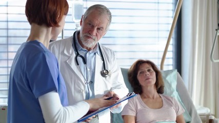 Wall Mural - Portrait of senior male doctor standing in hospital room, talking to a nurse.