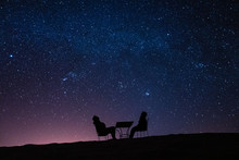 Young Couple Sitting At A Table On A Desert Dune While Talking, Relaxing And Observing The Stars And The Milky Way Above Them