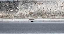 Grunge Weathered Concrete Wall With Exposed Stones And Bricks. Cement Sidewalk And Asphalt Road In Front. Background For Copy Space