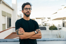 Confident Stylish Guy With Tattoos Posing On Apartment Balcony Or Terrace. Young Man In Glasses Standing Outside With Arms Crossed And Looking At Camera. Male Portrait Concept