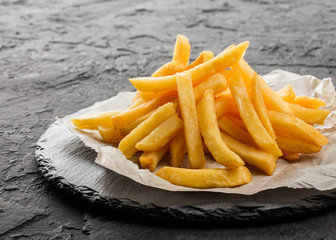 Canvas Print - Tasty french fries potatoes on paper over black stone background. Hot fast food, close up