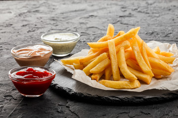 Canvas Print - Tasty french fries potatoes on paper with sauces over black stone background. Hot fast food