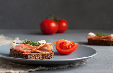 A grey plate with black bread and bacon, a sprig of dill, and a sliced tomato. In the background, two tomatoes and a sandwich. On a grey background