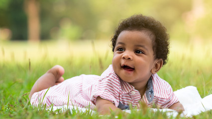 Cute little dark skinned baby boy smiling with happiness while lying on the grass in the park.