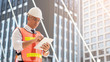 © Dollydoll - Caucasian engineering man wearing white hard hat while checking on the tablet at the work side with building all around.