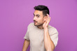 Young handsome man over isolated purple background listening to something by putting hand on the ear
