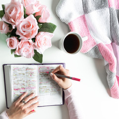 Wall Mural - Pink, white, grey Bible flat lay with black tea, pink roses, colorful Bible, pen, coffee and a woman’s hand writing in a journal. Prayer time, quiet time with God, morning devotional