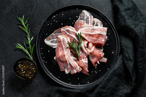 Cuadros en Lienzo Raw bacon on a black plate, pepper and rosemary