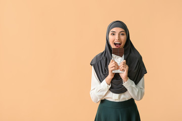 Beautiful Muslim woman eating tasty chocolate on color background